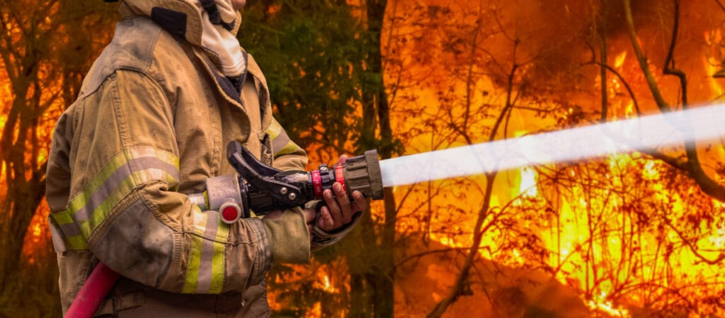 Firefighter in protective gear spraying water to combat intense wildfire in forest, representing emergency response and wildfire suppression efforts